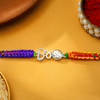 A braided Rakhi bracelet with a silver Om symbol and a silver 'B' letter charm, featuring purple on one side and orange-red on the other, laid out on a yellow surface.